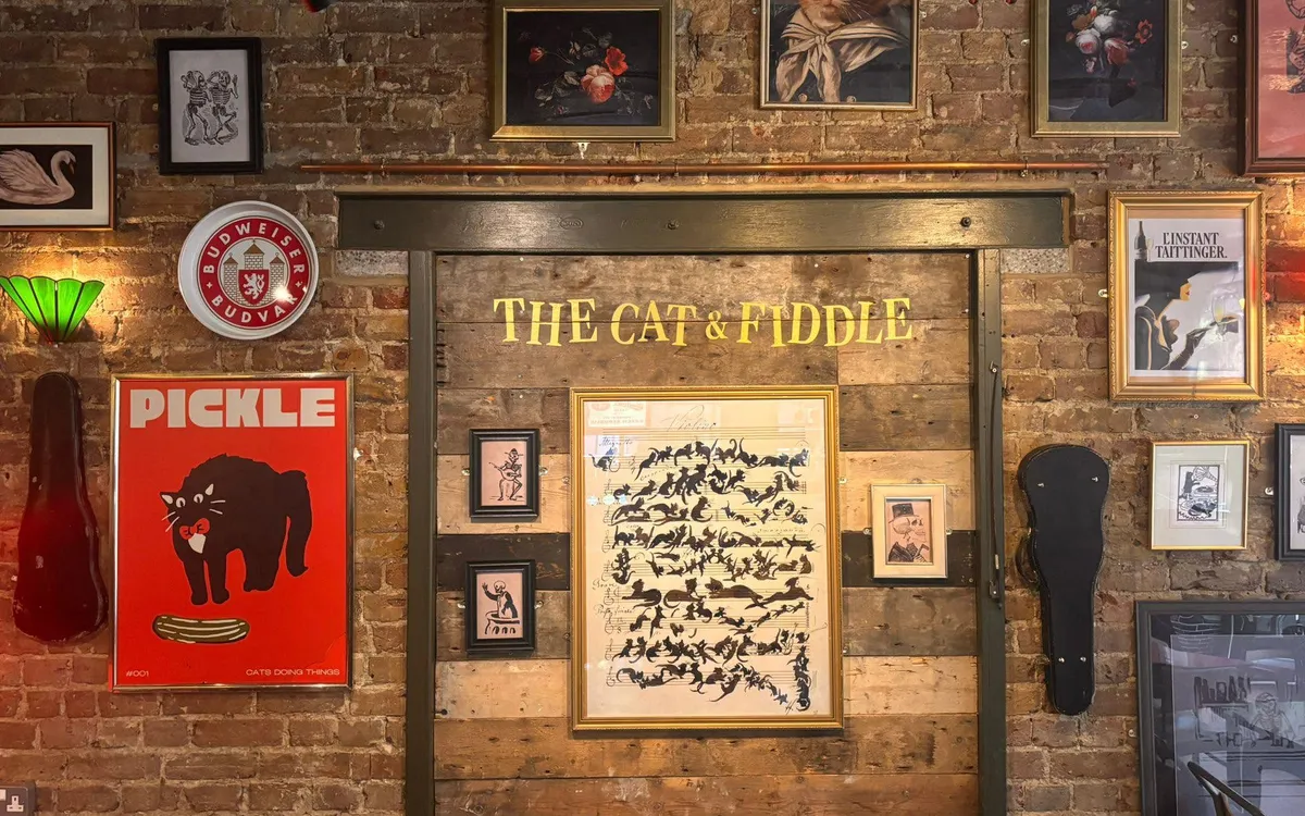 The Cat & Fiddle’s brick back wall, covered in framed prints and gig posters, with its name painted in gold
