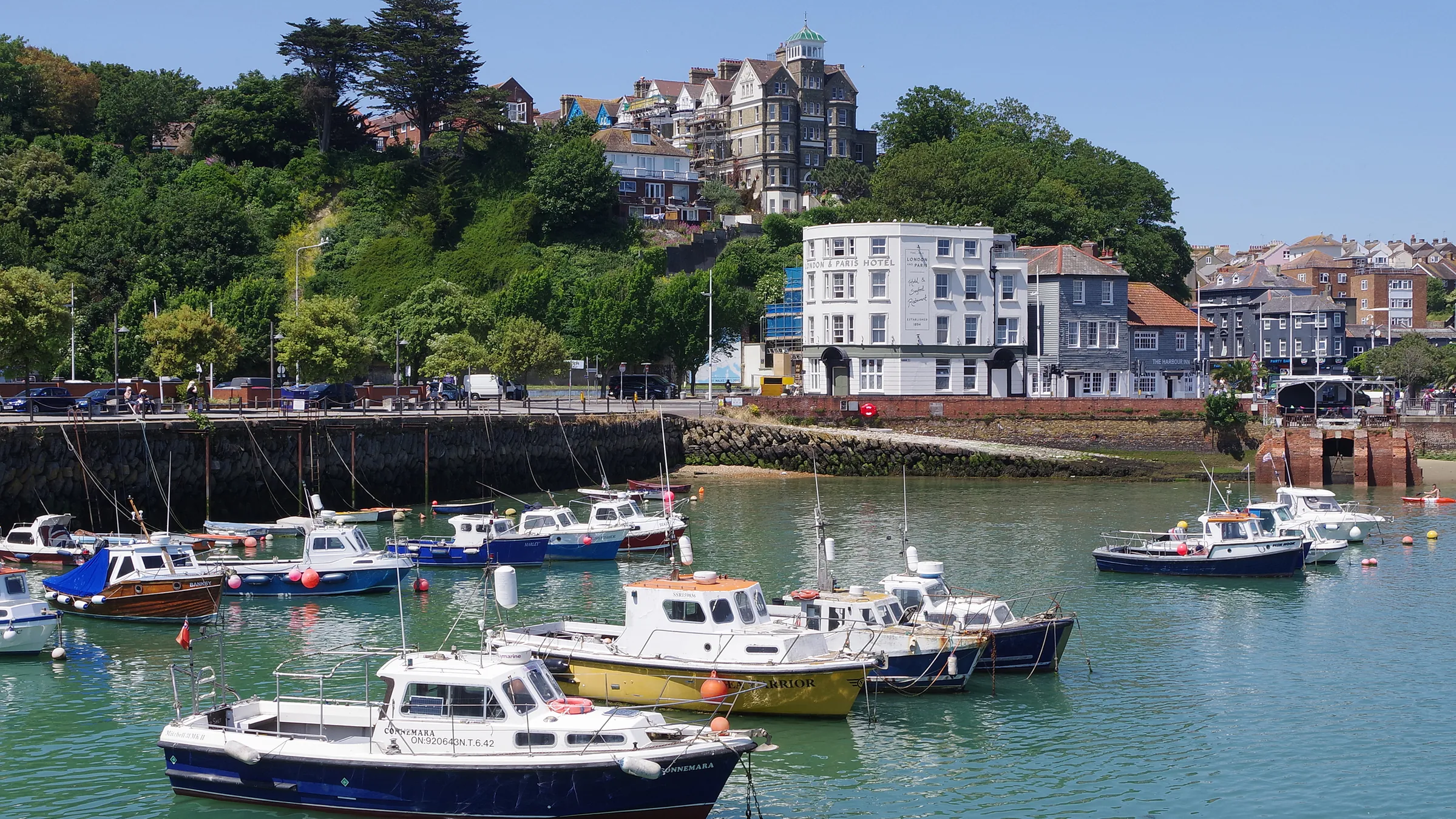 Colourful boats at high tide in Folkestone Harbour, with the white London & Paris Hotel on the waterfront and wooded cliffs behind